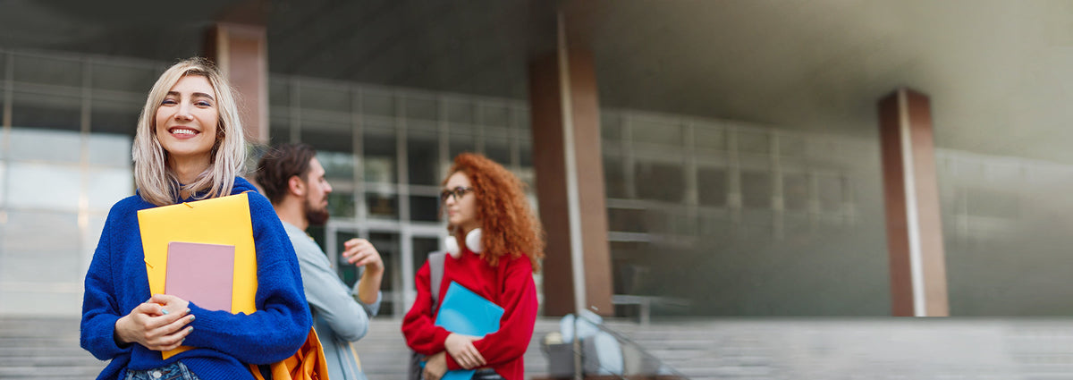 Woman holding books in front of a university with other people in the background.