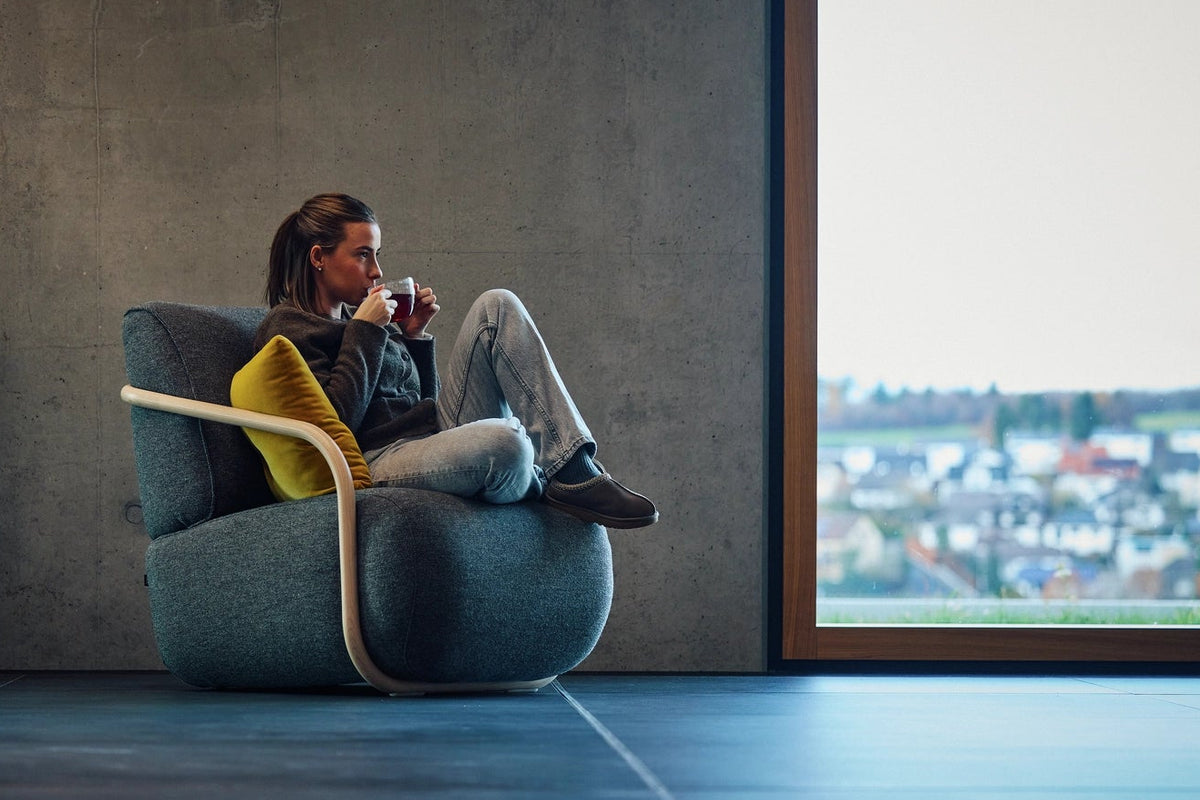 A woman sits in a modern gray lounge chair, drinking from a mug, in a minimalist concrete room with a large window.