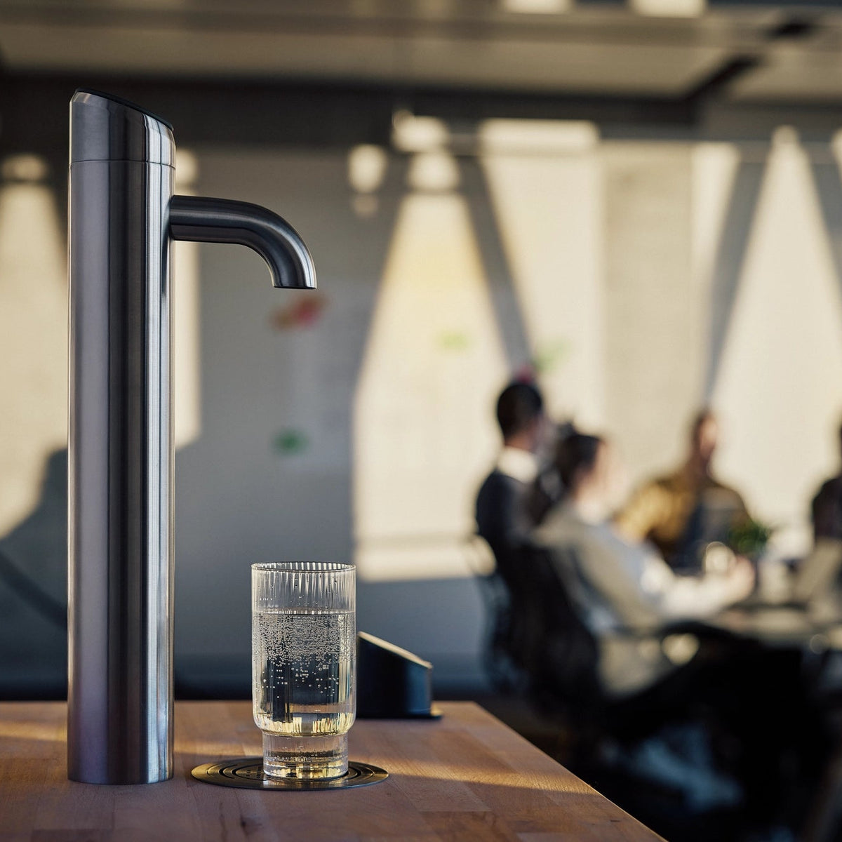 BRITA Extra I-Tap water dispenser on a wooden table with a glass of water underneath in a blurred office setting.